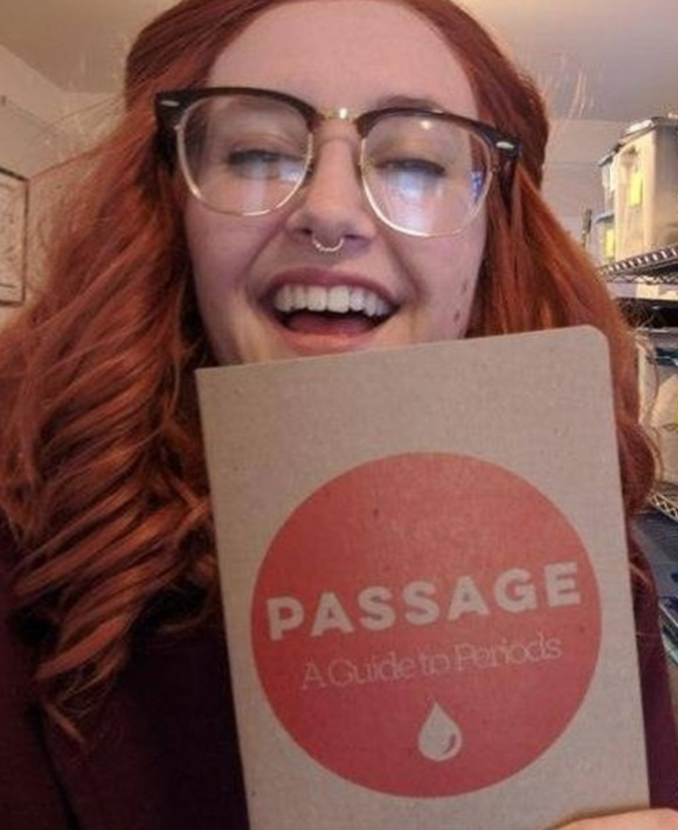 A smiling Kelsey in glasses holds up the new print copy of Passage: A Guide To Periods.