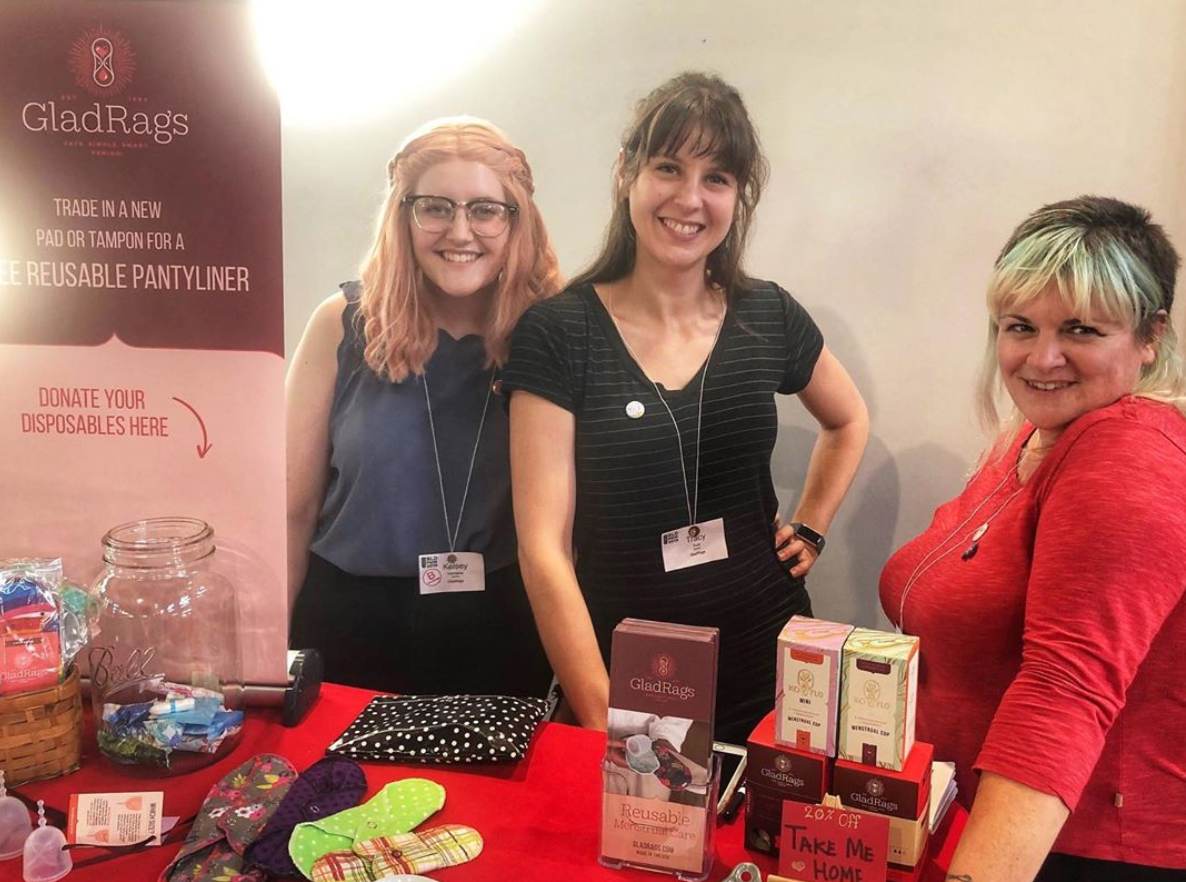 Kelsey, Tracy, and Margaux of GladRags volunteer with Project Lemonade for B Corp Day Of Service. They're standing at a table with GladRags products.