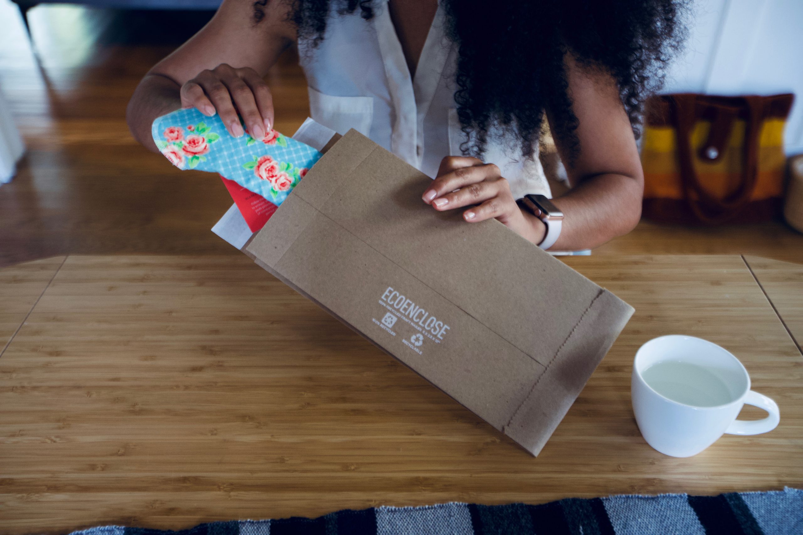 A person sitting at a table pulls an unwrapped cloth pad out of a brown eco mailer.