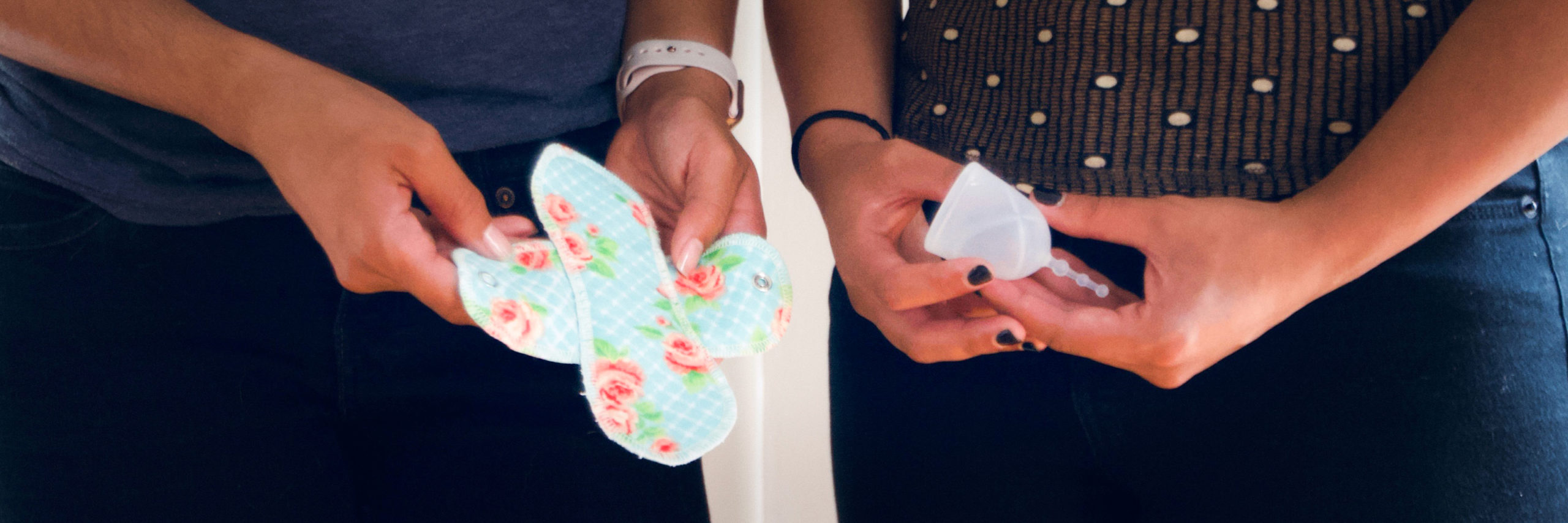 Two people each hold a reusable menstrual product, one holds a cloth pad with a pink and green floral print on a blue background, the other holds a menstrual cup.