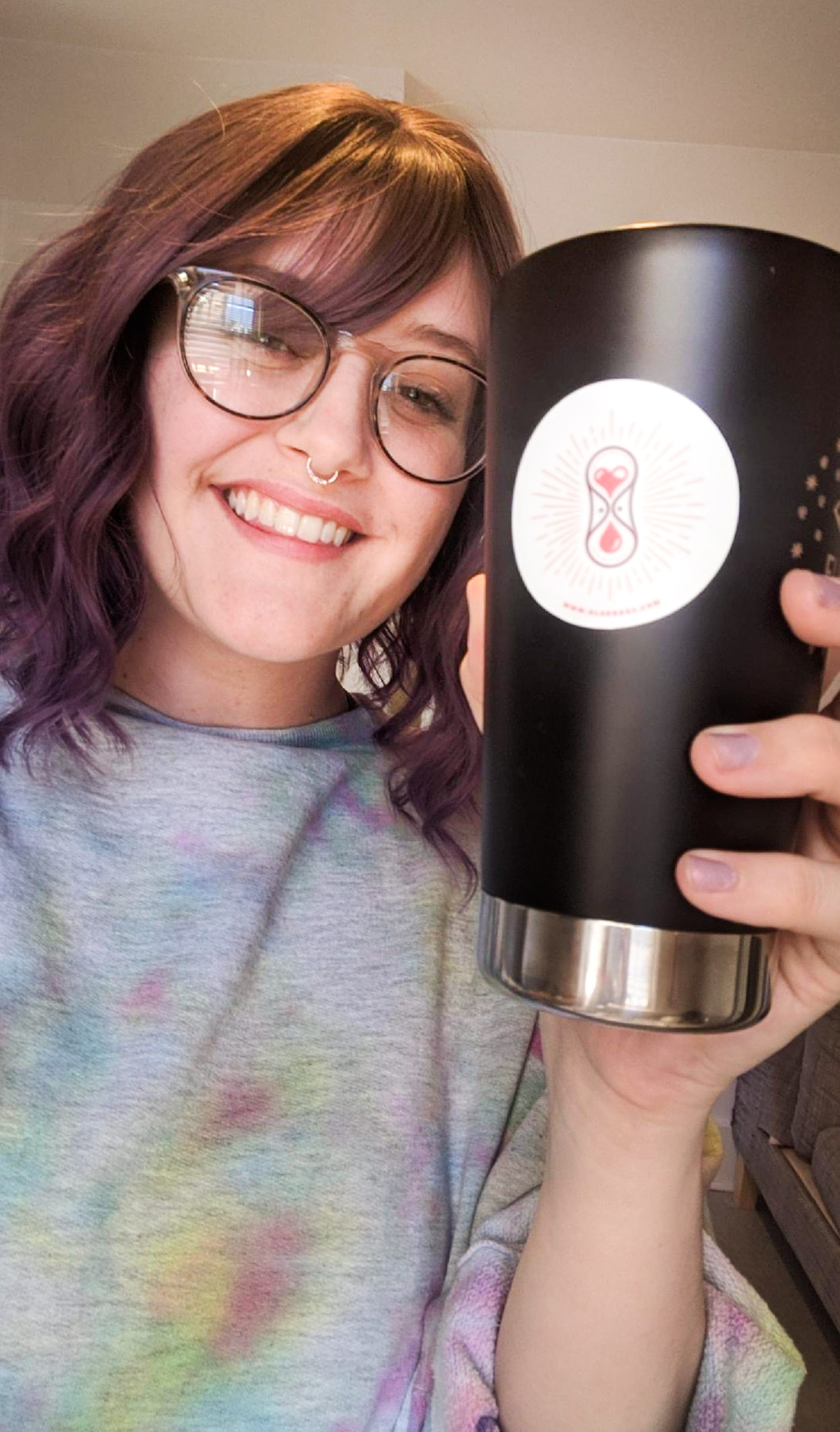 A smiling person with a great smile and wearing glasses holds out a reusable mug in a cheers pose.