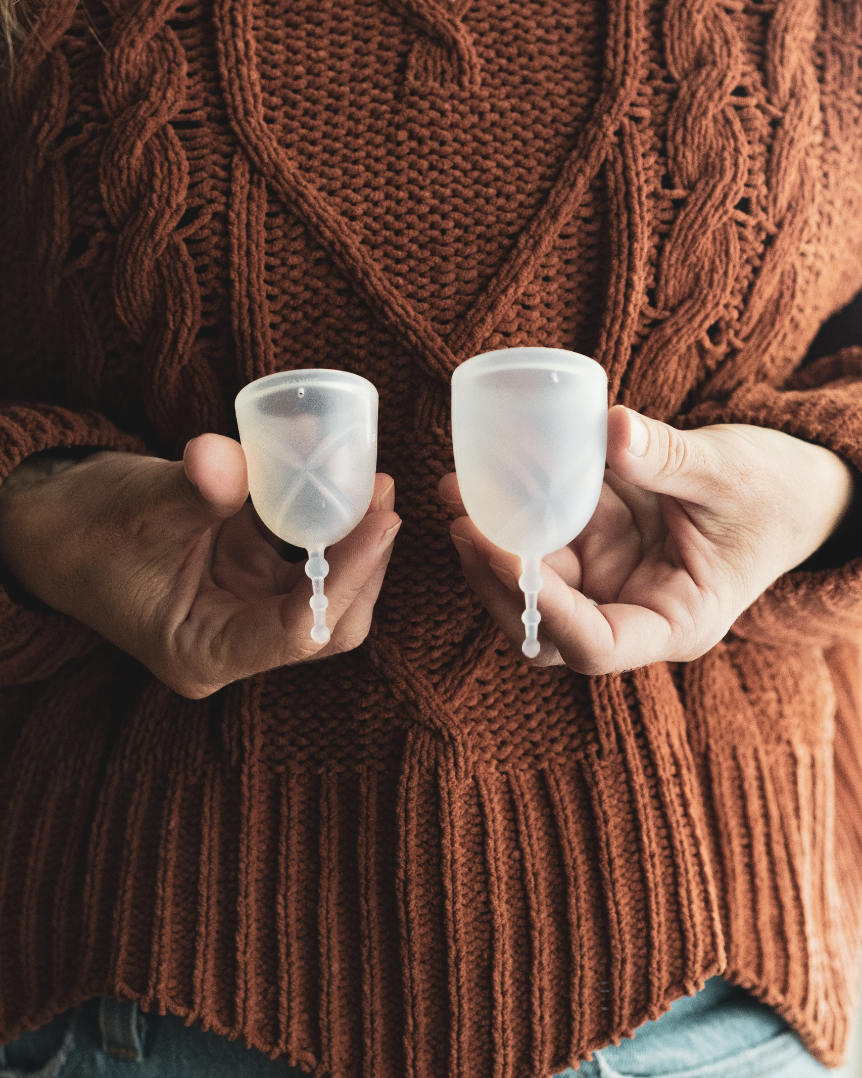 A person with an orange sweater holds two menstrual cups in front of their body.