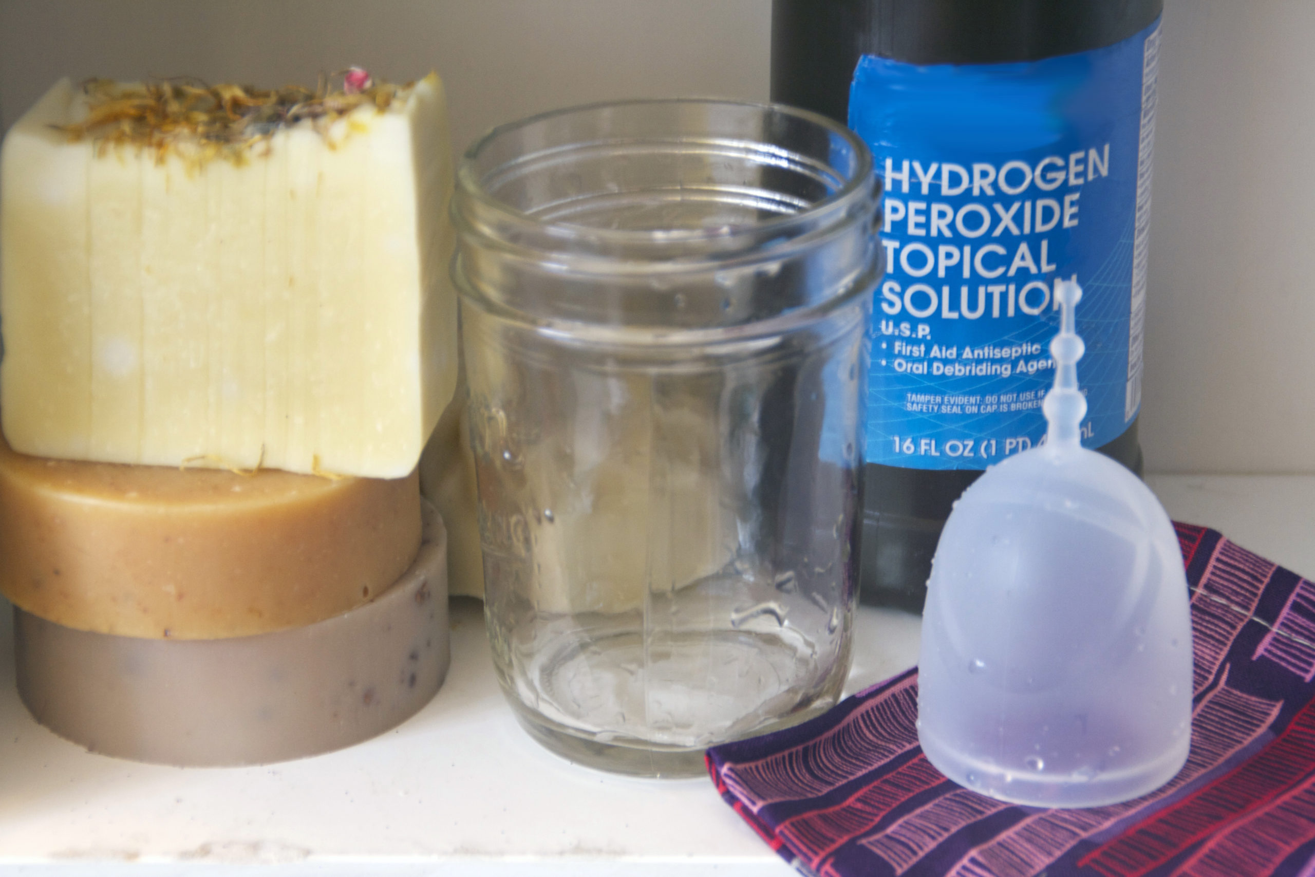 A clear glass jar sits on a shelf next to a bottle of hydrogen peroxide and a menstrual cup. 