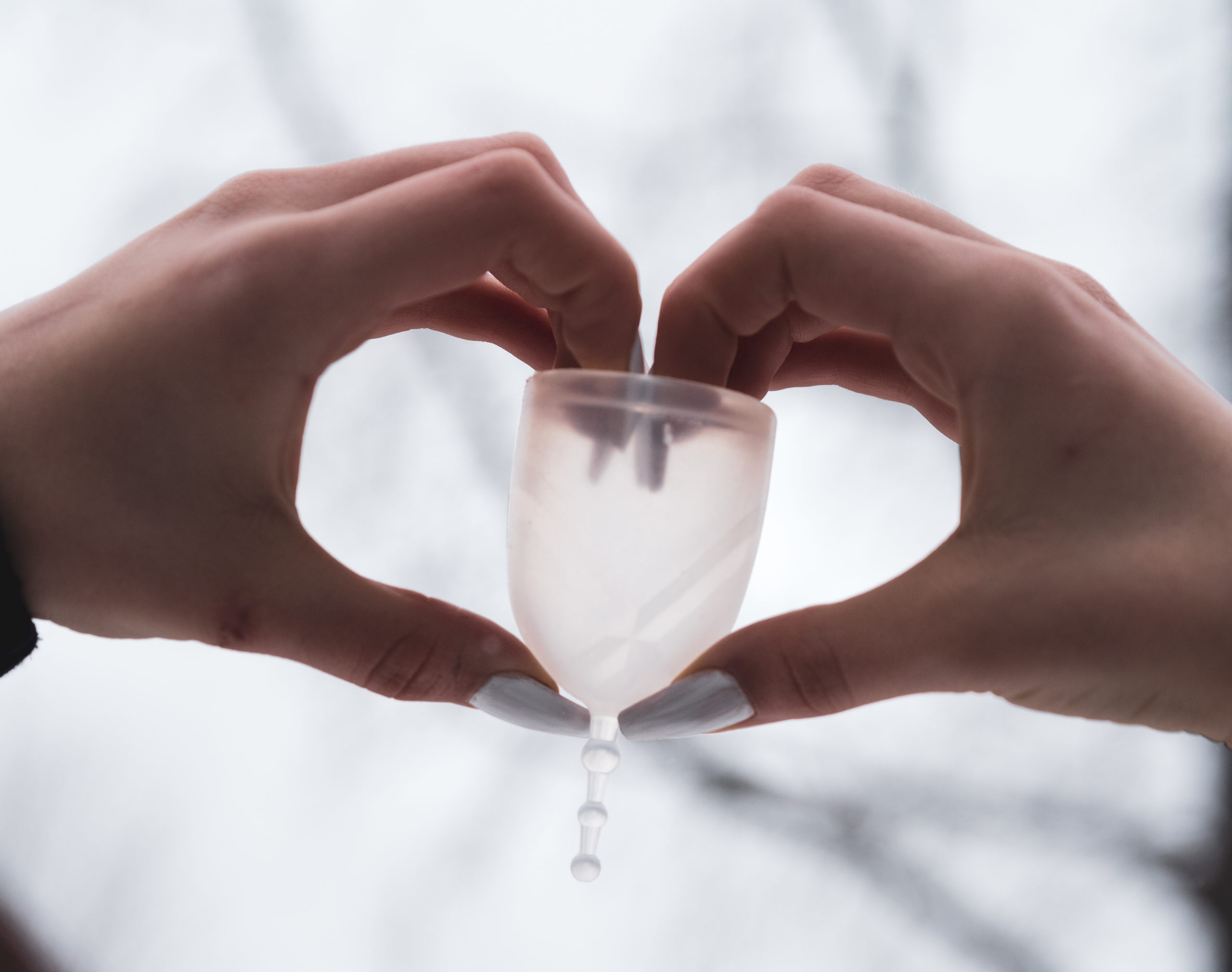 A pair of hands with long silver nails forms the shape of a heart and holds up a clear menstrual cup.