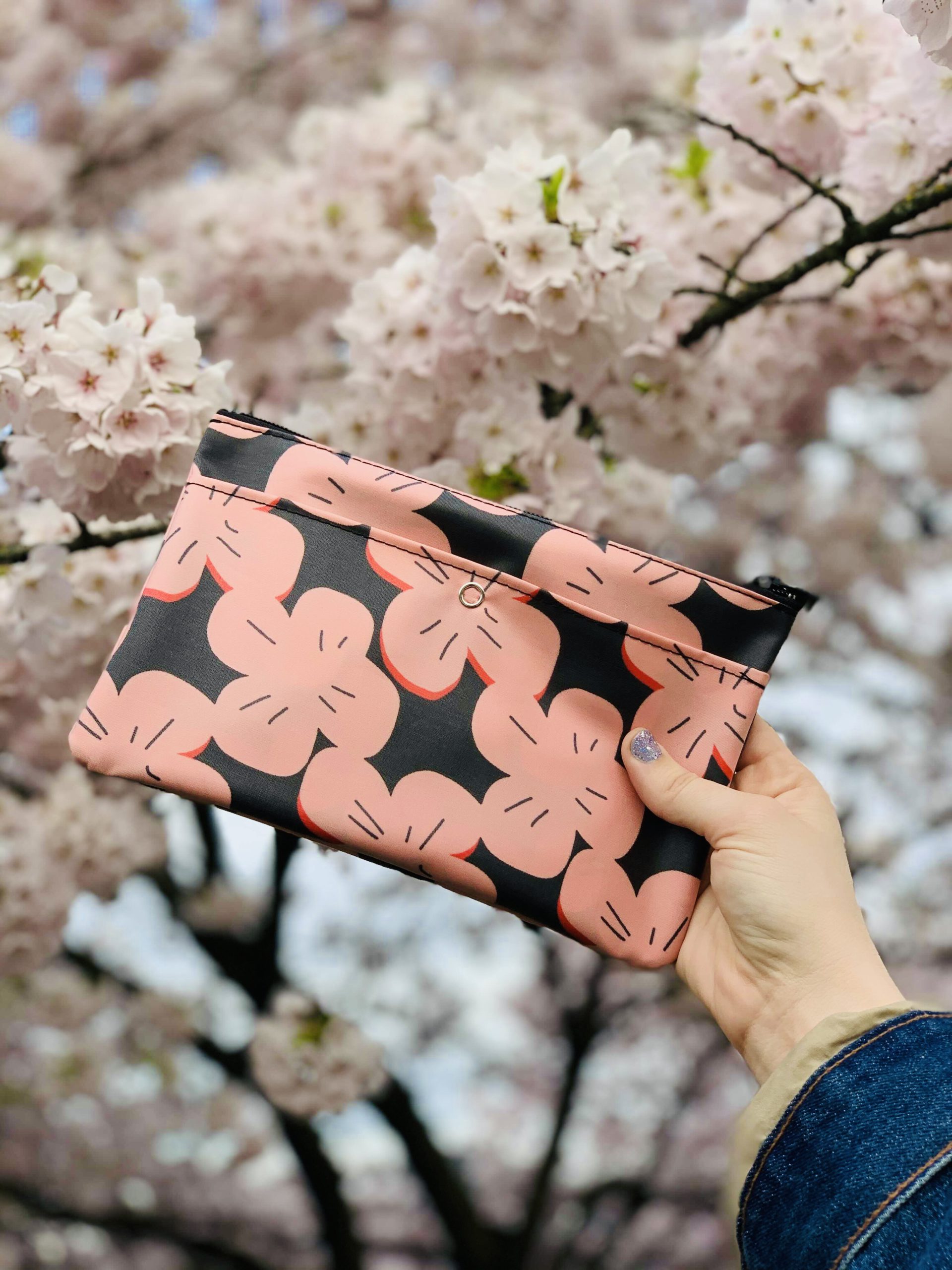 A fair-skinned hand with purple fingernails holds up a waterproof carry bag in a cherry blossom print in front of real cherry blossoms on a tree. 