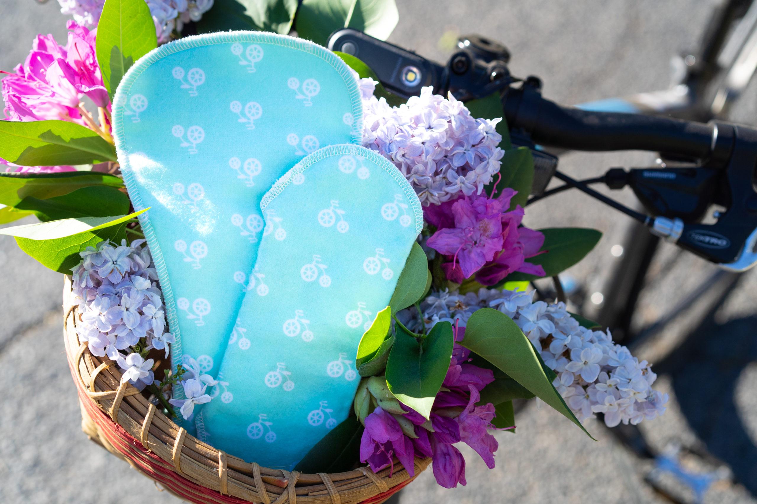 Two cloth pads in a mint green with a white bicycle print are in a bicycle basket with purple lilacs and pink rhododendron blooms.