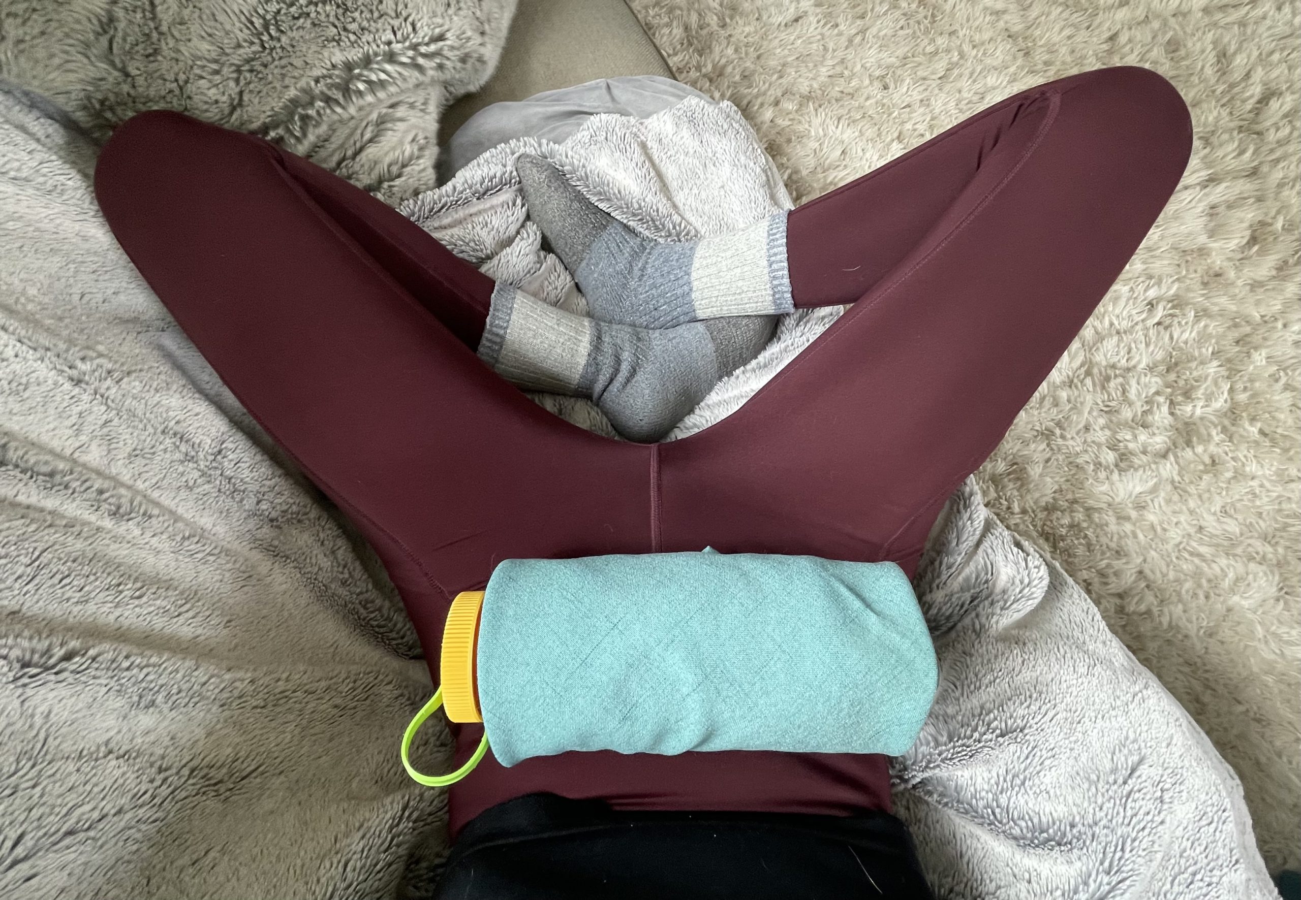 The point of view looking down into a lap, a pair of legs bent at the knees, with gray socks on the feet, and a cloth napkin wrapped around a yellow-lidded water bottle against the body's lower abdomen.