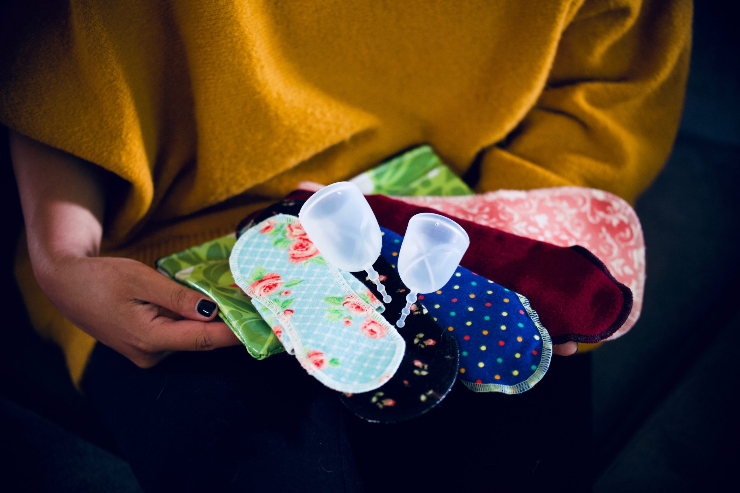 A person in a yellow sweater holds an array of cloth pads in various prints, and two translucent menstrual cups. 