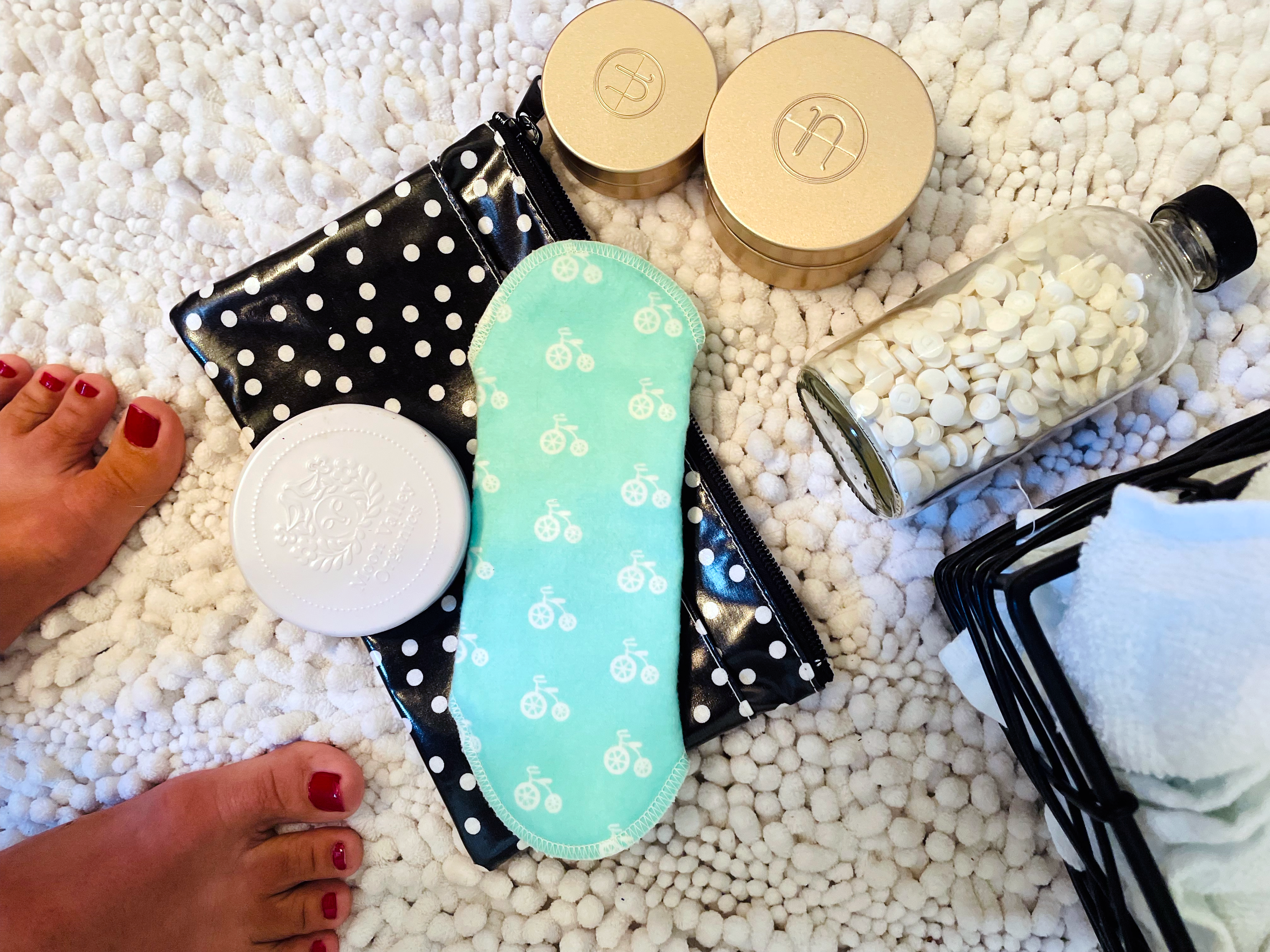 An overhead shot of plastic free personal care items on a fluffy white bath rug, including a turquoise cloth pad with a bicycle print, toothpaste bites, and shampoo bars. 